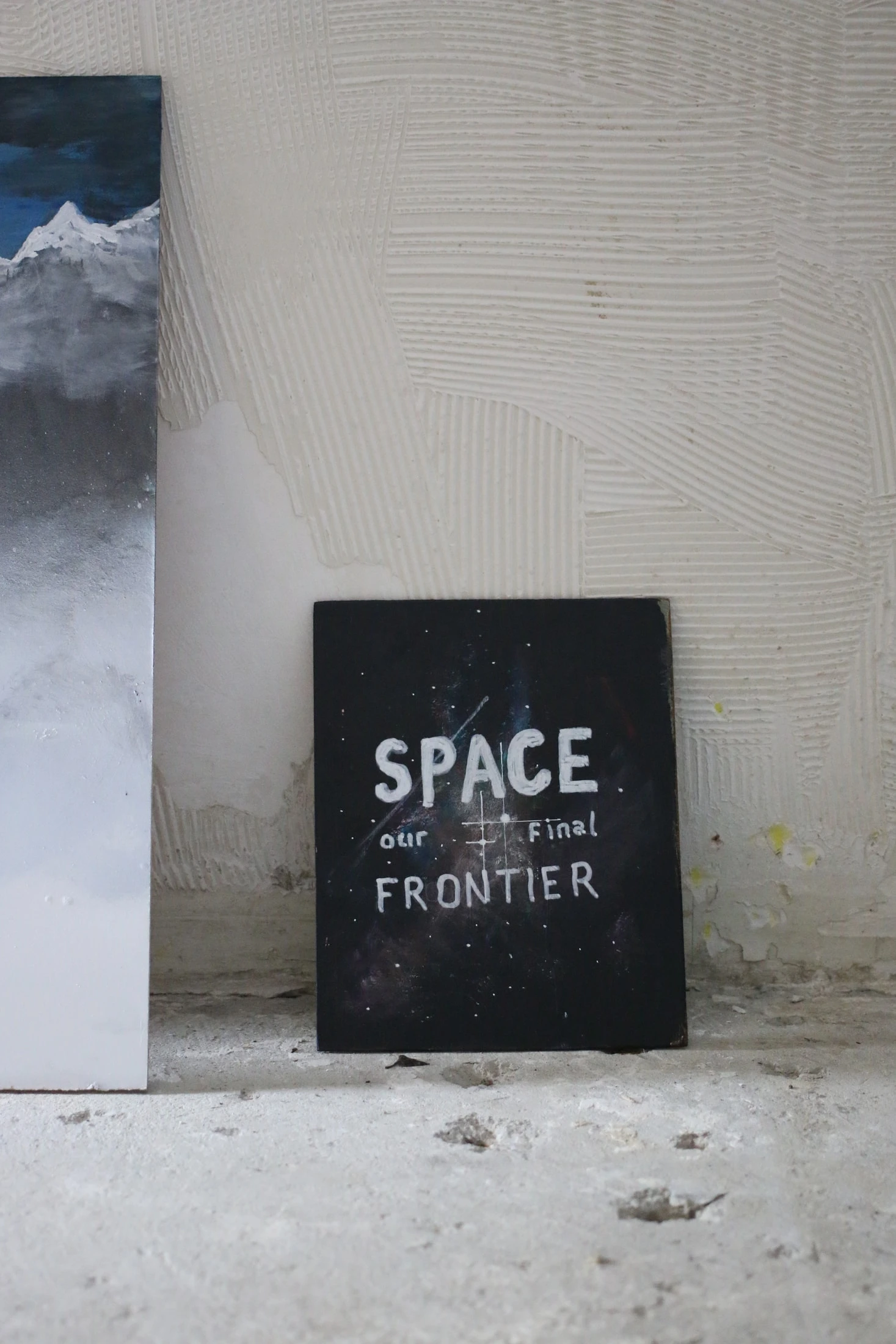A dark painting with white lettering reading SPACE our Final FRONTIER, leaning against a studio wall.