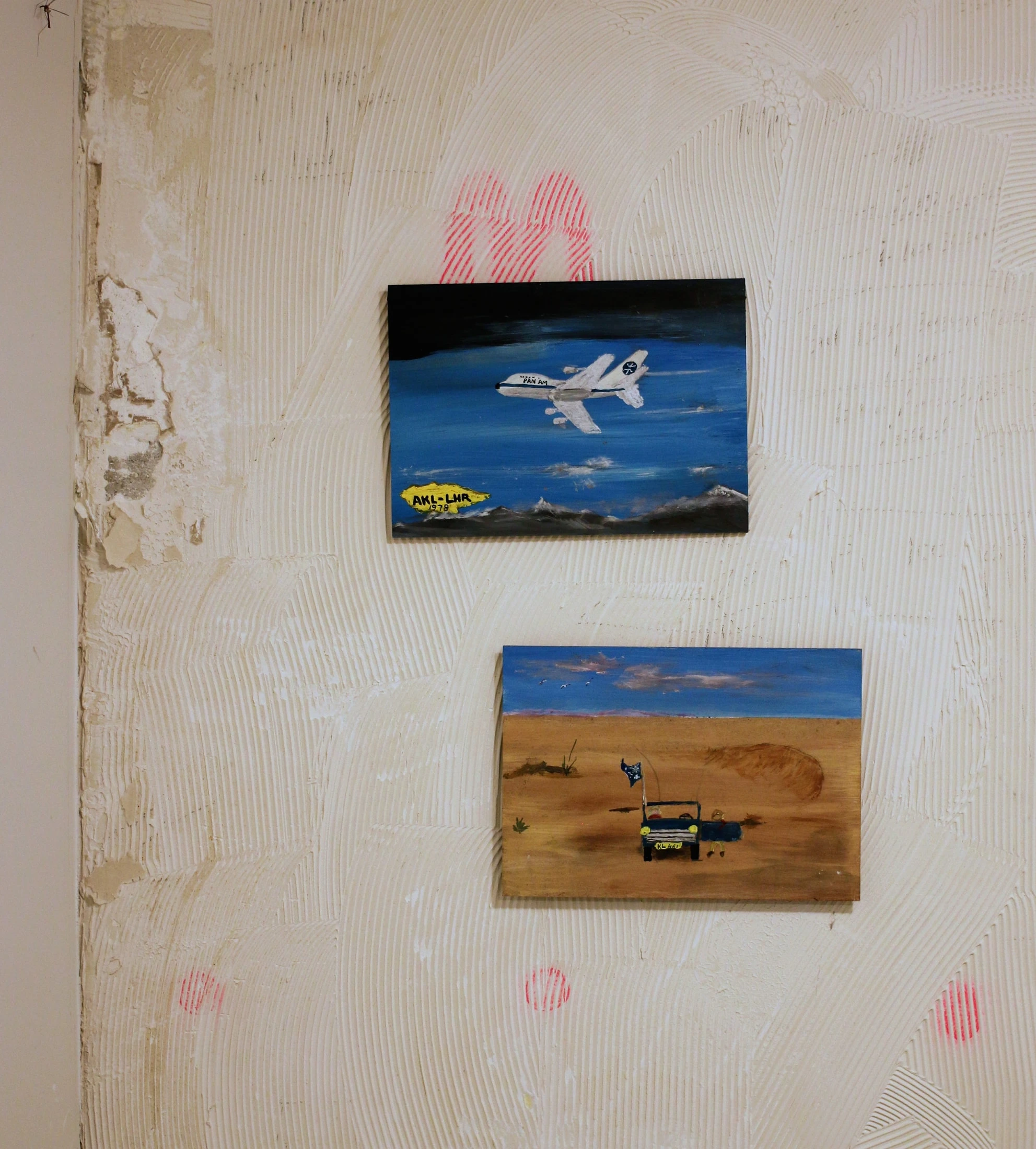 Two paintings on a studio wall: a white passenger aircraft above mountains at the top, and a vehicle in a desert below.